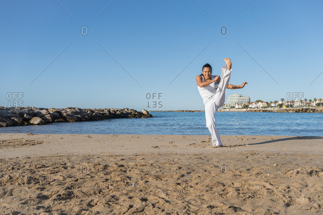Young focused ethnic female in capoeira clothes showing balance trick while standing with raised arms on sandy shore near ocean and looking at camera