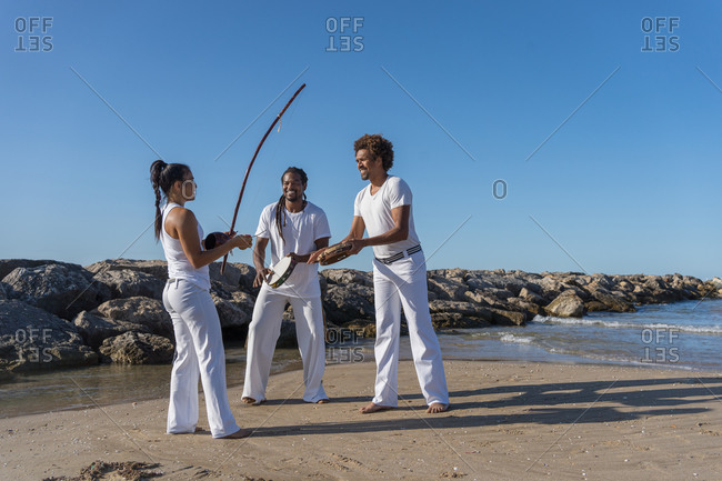 Cheerful multicultural group of people playing musical instruments near woman with archery bow during capoeira performance under blue sky
