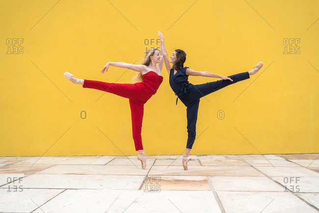 Full body side view of talented young female dancers in colorful outfits performing dancing movement with legs raised against yellow wall