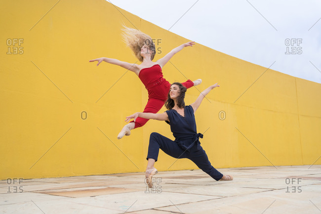 Full body of talented young female dancers in stylish colorful clothes jumping and performing graceful dynamic movement near yellow wall on street