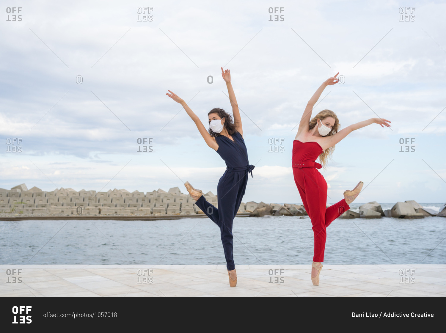 Full body of slim young women in colorful clothing standing on tiptoe and performing graceful dance on paved embankment against cloudy sky