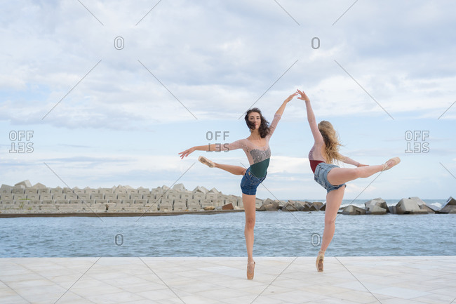 Full body of young slim female dancers standing on tiptoe in graceful dance position on paved embankment near sea
