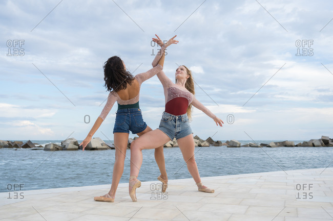 Full body of professional slim female ballet dancers performing contemporary active dance on paved seafront against cloudy sky