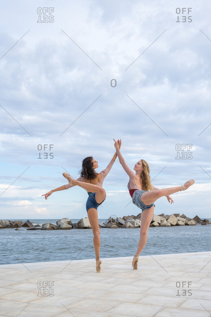 Full body side view of elegant professional modern ballet dancers balancing on tiptoe while performing sensual dance near sea against cloudy sky