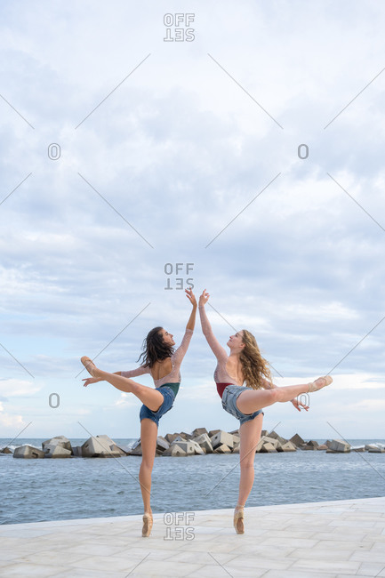 Full body side view of elegant professional modern ballet dancers balancing on tiptoe while performing sensual dance near sea against cloudy sky