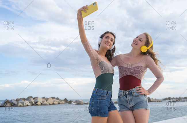 Joyful young girlfriends taking selfie with smartphone while having fun and listening to music with headphones on seashore