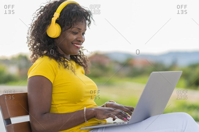 Side view of delighted african american self employed woman in headphones sitting on bench