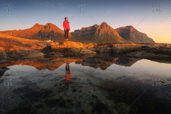 Unrecognizable female tourist admiring highland while standing on mountain near pure pond under sky at sunset
