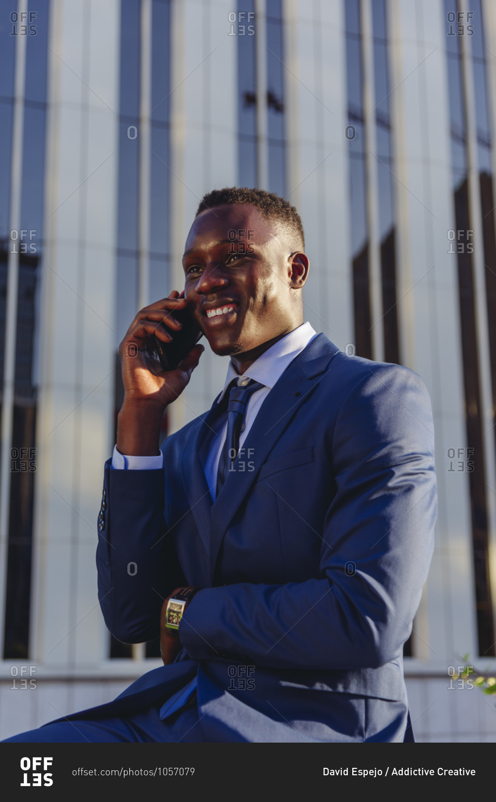 Low angle of happy successful well dressed black male manager in classy suit answering phone call while sitting against contemporary city building