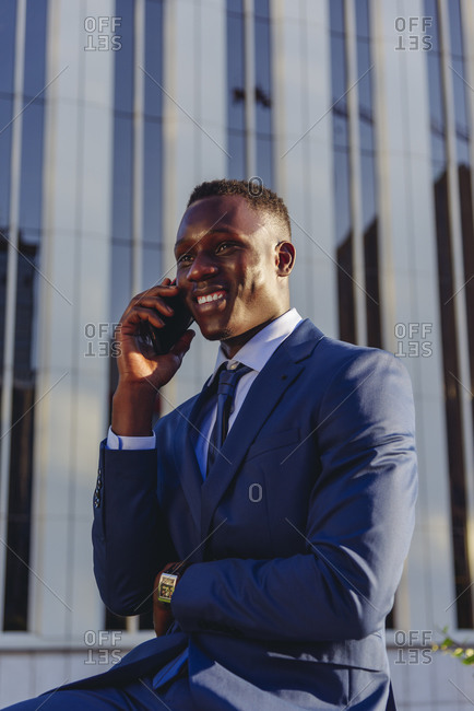 Low angle of happy successful well dressed black male manager in classy suit answering phone call while sitting against contemporary city building
