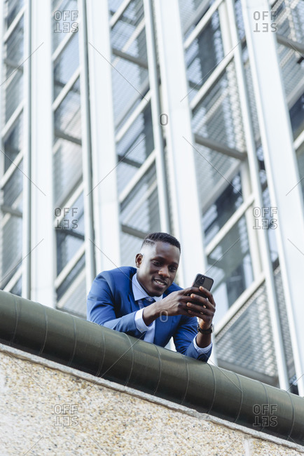 Low angle of confident respectable african american businessman in formal outfit leaning on street against contemporary buildings in modern downtown with smartphone