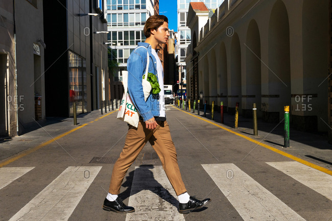 Full body side view of handsome young male in stylish outfit with reusable textile bag with groceries crossing road in modern city district
