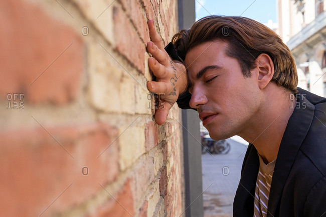 Side view of young reflective trendy male with closed eyes standing leaned with hand on shabby wall on street