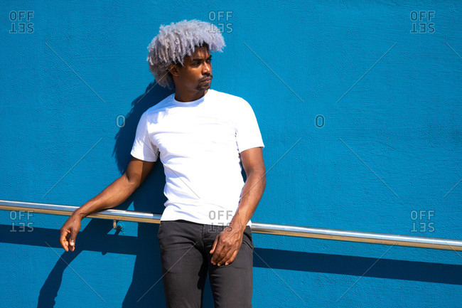 Black man with afro hair dressed in white leaning against a blue wall. afro hair concept. black man in white t-shirt.