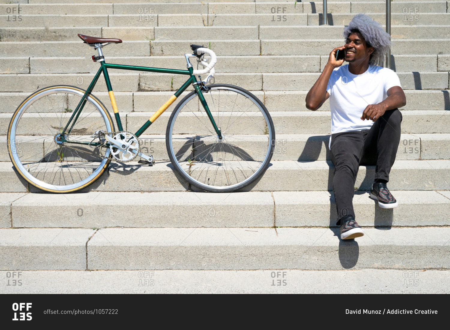 Black man talking on the phone next to his bike. man sitting on the stairs and calling with his cell phone.