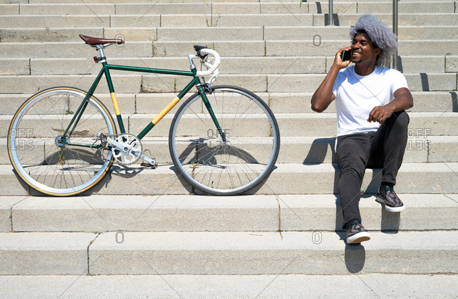 Black man talking on the phone next to his bike. man sitting on the stairs and calling with his cell phone.
