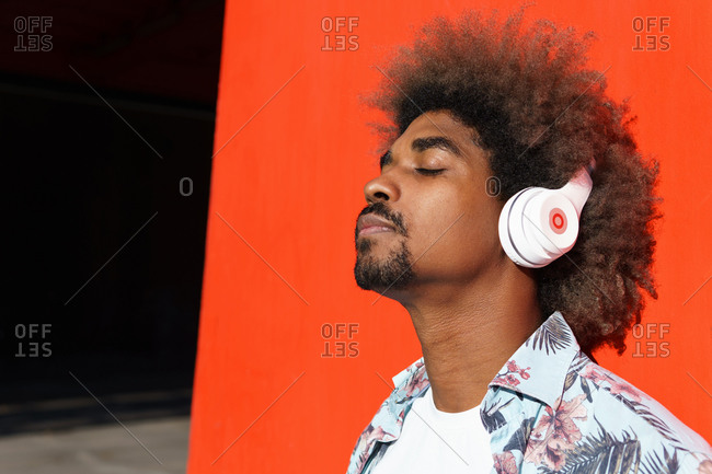 Young masculine bearded african american male with afro hairstyle and closed eyes listening to music in headphones near red wall in town