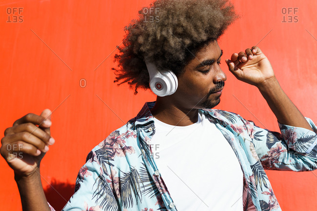 Young masculine bearded african american male with afro hairstyle and closed eyes listening to music in headphones near red wall in town