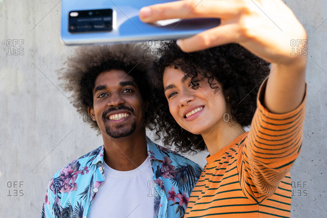 Content multiracial couple of friends with afro hairstyle taking selfie on cellphone while embracing near wall in sunlight in back lit