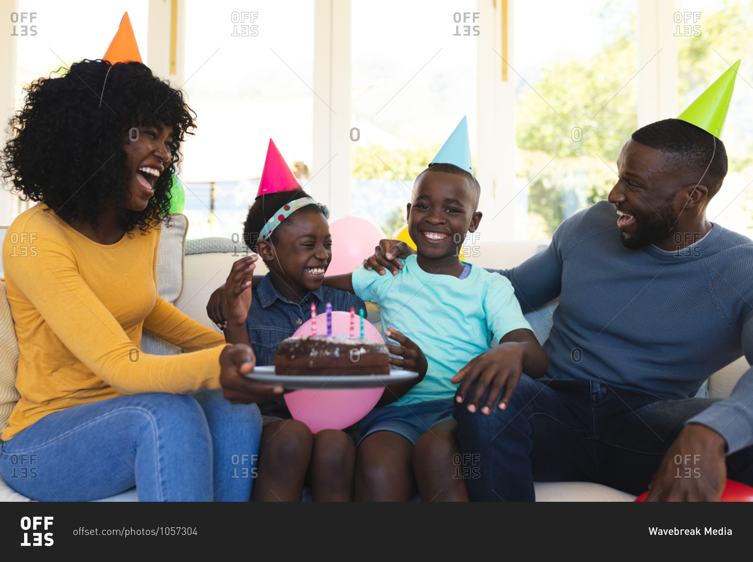 African American family wearing party hats with cake celebrating birthday sitting on the couch in the living room at home..