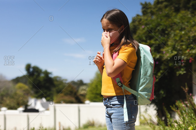 Caucasian girl with dark hair wearing face mask walking to school carrying schoolbag.