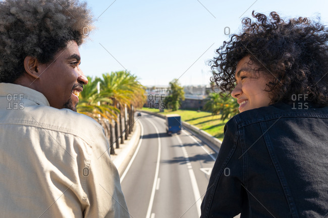 Back view of happy young ethnic woman and man smiling and looking at each other while standing on roadside on sunny day