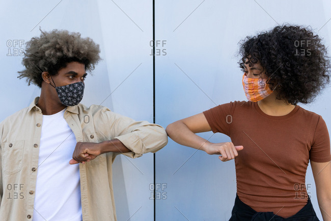 Anonymous young african american man and woman with curly hairs in casual clothes and protective masks touching elbows against white wall