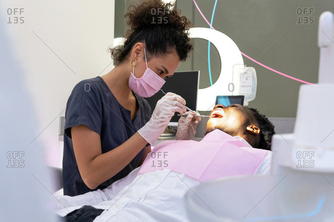 African american female doctor in mask and gloves doing dental procedure with mirror and metal explorer for patient lying under lamp in medical room