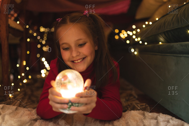 Caucasian girl smiling and holding snow globe, lying in blanket fort.