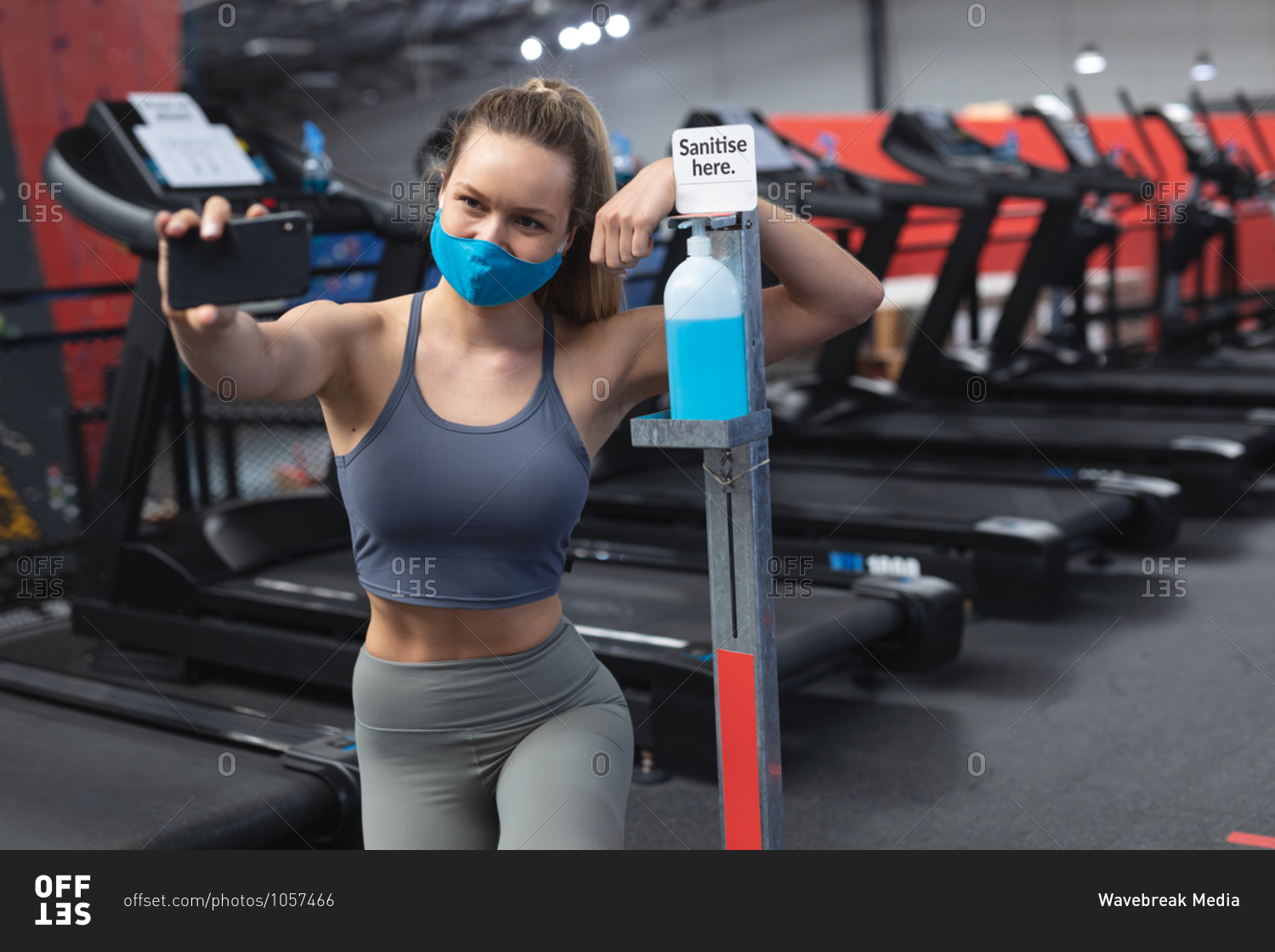 Fit caucasian woman wearing face mask taking a selfie with sanitizer foot dispenser stand with sanitize here text in the gym.