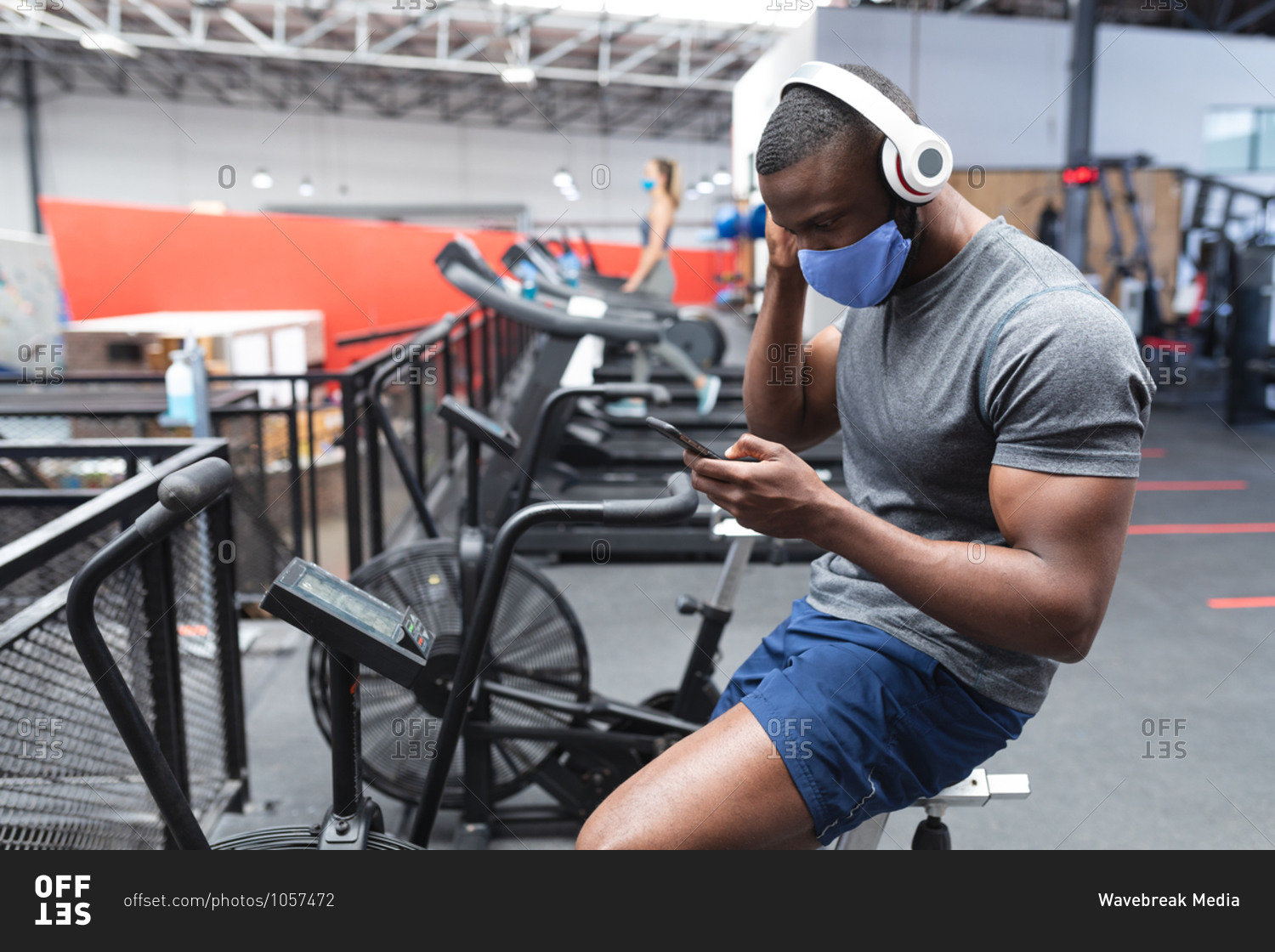 Fit African American man wearing face mask and headphones using smartphone while sitting on stationary bike in the gym.