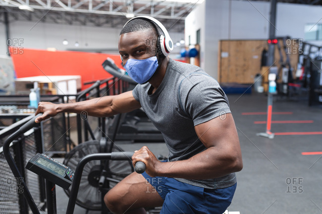 Portrait of fit African American man wearing face mask and headphones exercising on stationary bike in the gym.