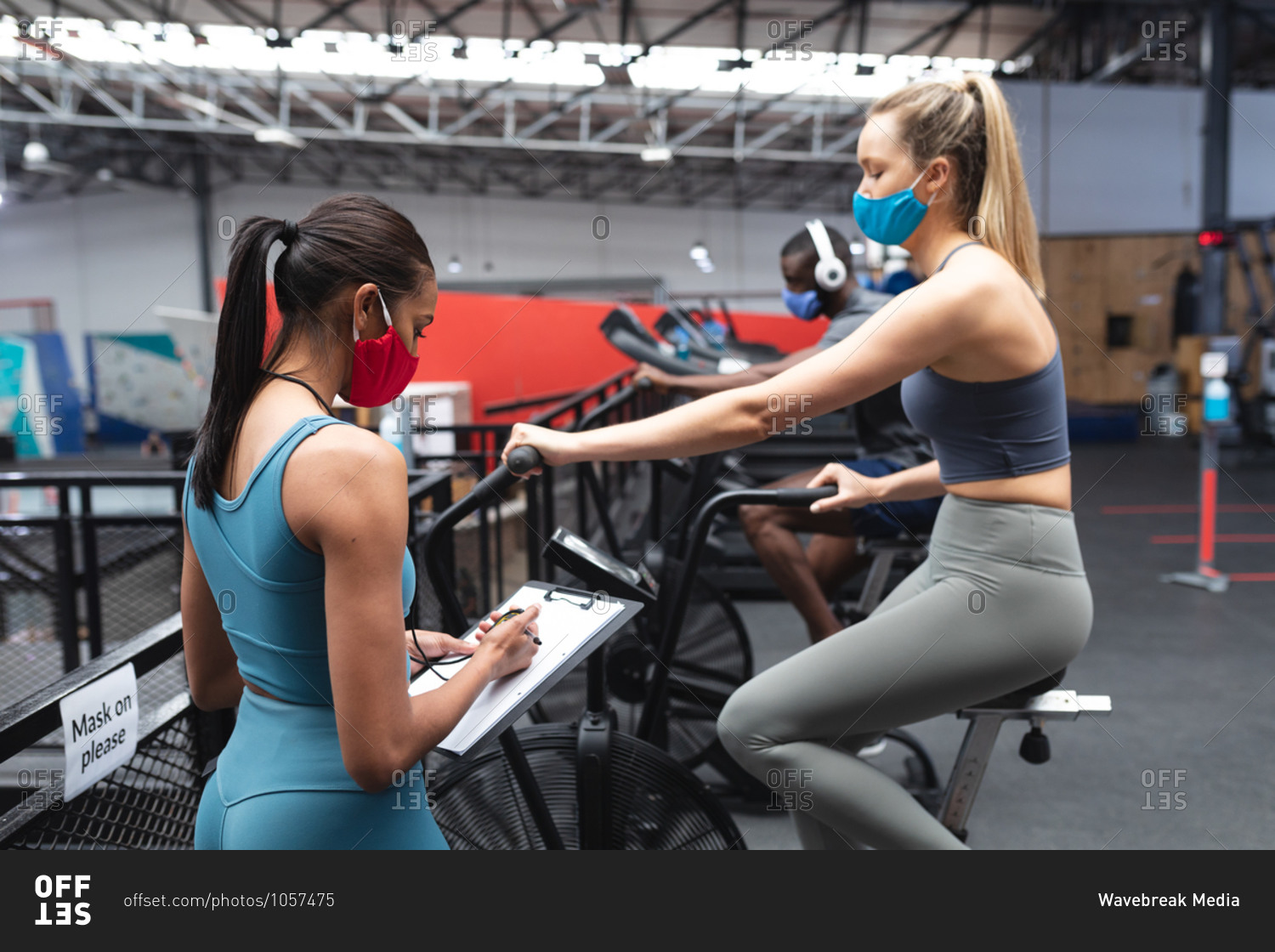 Fit caucasian woman wearing face mask exercising on stationary bike while caucasian female fitness coach taking notes on clipboard in the gym.
