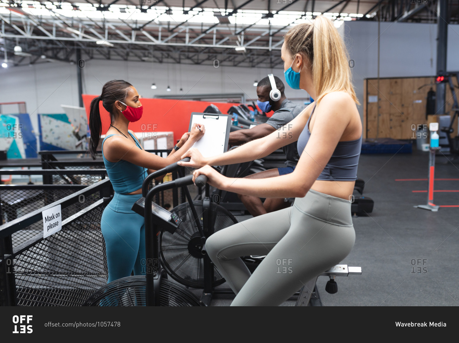 Fit caucasian woman wearing face mask exercising on stationary bike while caucasian female fitness coach taking notes on clipboard in the gym.