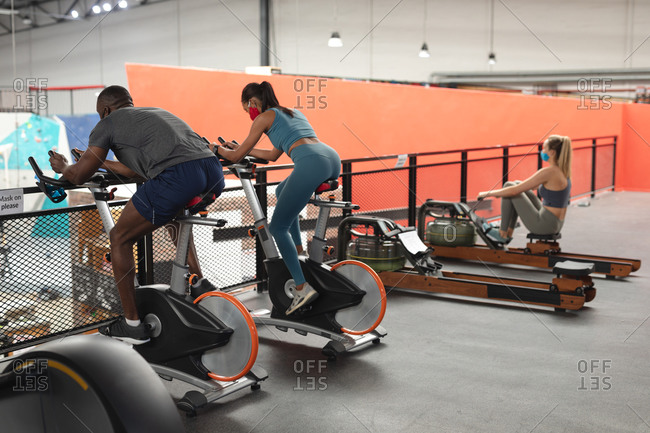 Fit African American man and fit caucasian woman wearing face masks exercising on stationary bike in the gym.