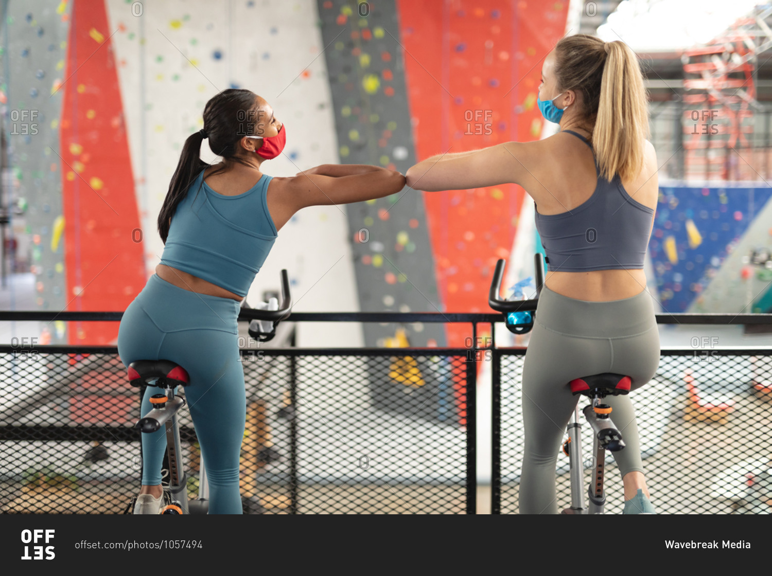Rear view of two fit caucasian woman wearing face masks greeting each other by touching elbows while exercising on stationary bike in the gym.