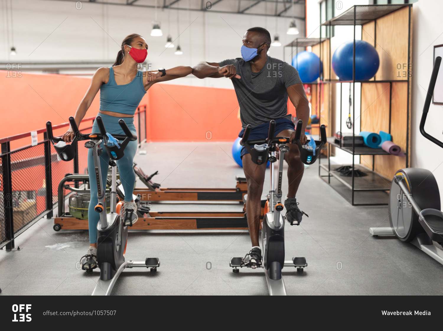 Fit African American man and fit caucasian woman wearing face masks greeting each other by touching elbows while exercising on stationary bike in the gym.