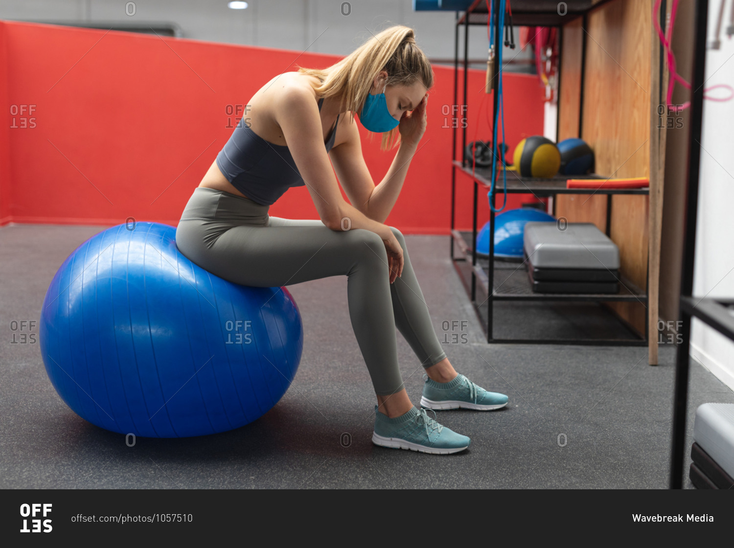 Stressed fit caucasian woman wearing face mask sitting on exercise ball in the gym.