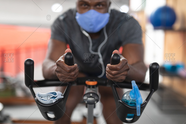 Portrait of fit African American man wearing face mask and earphones exercising on stationary bike in the gym.