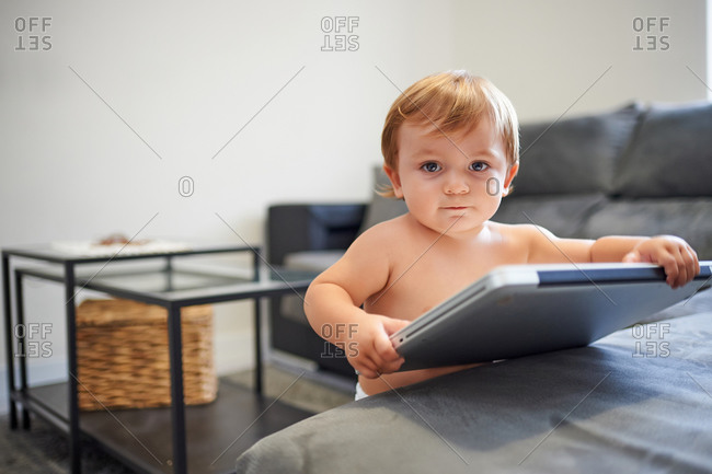 Adorable toddler with naked torso standing with netbook near couch at home and looking at camera