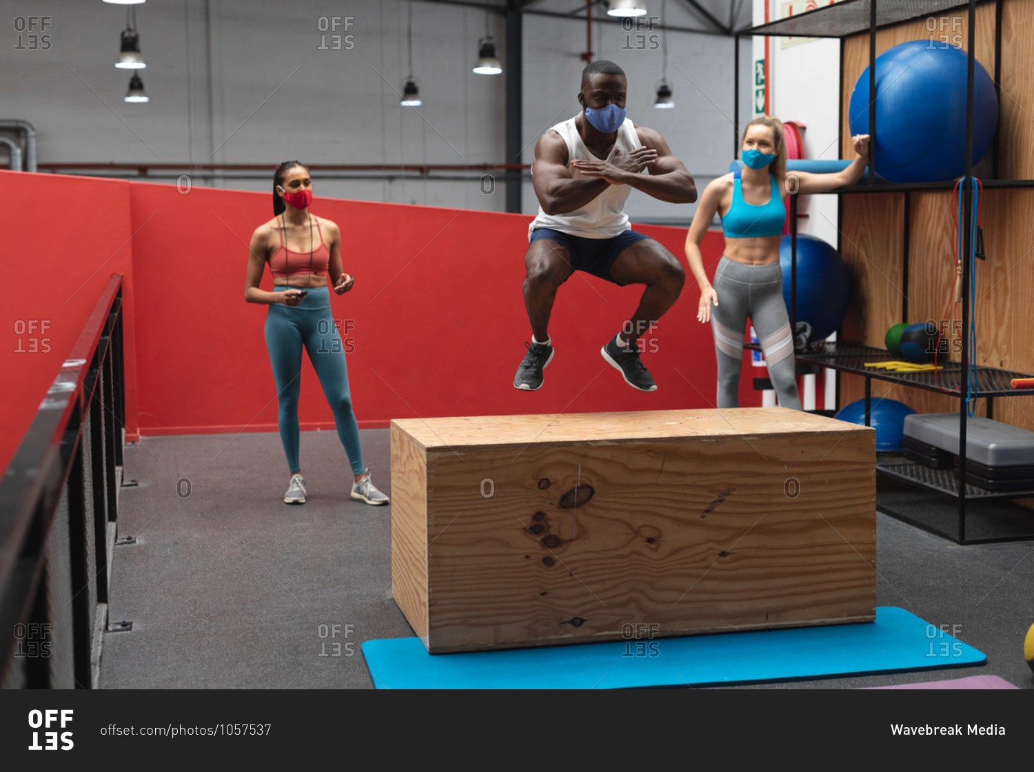 Fit African American man wearing face mask jumping on wooden plyo box in the gym while caucasian female fitness trainer holding stopwatch.