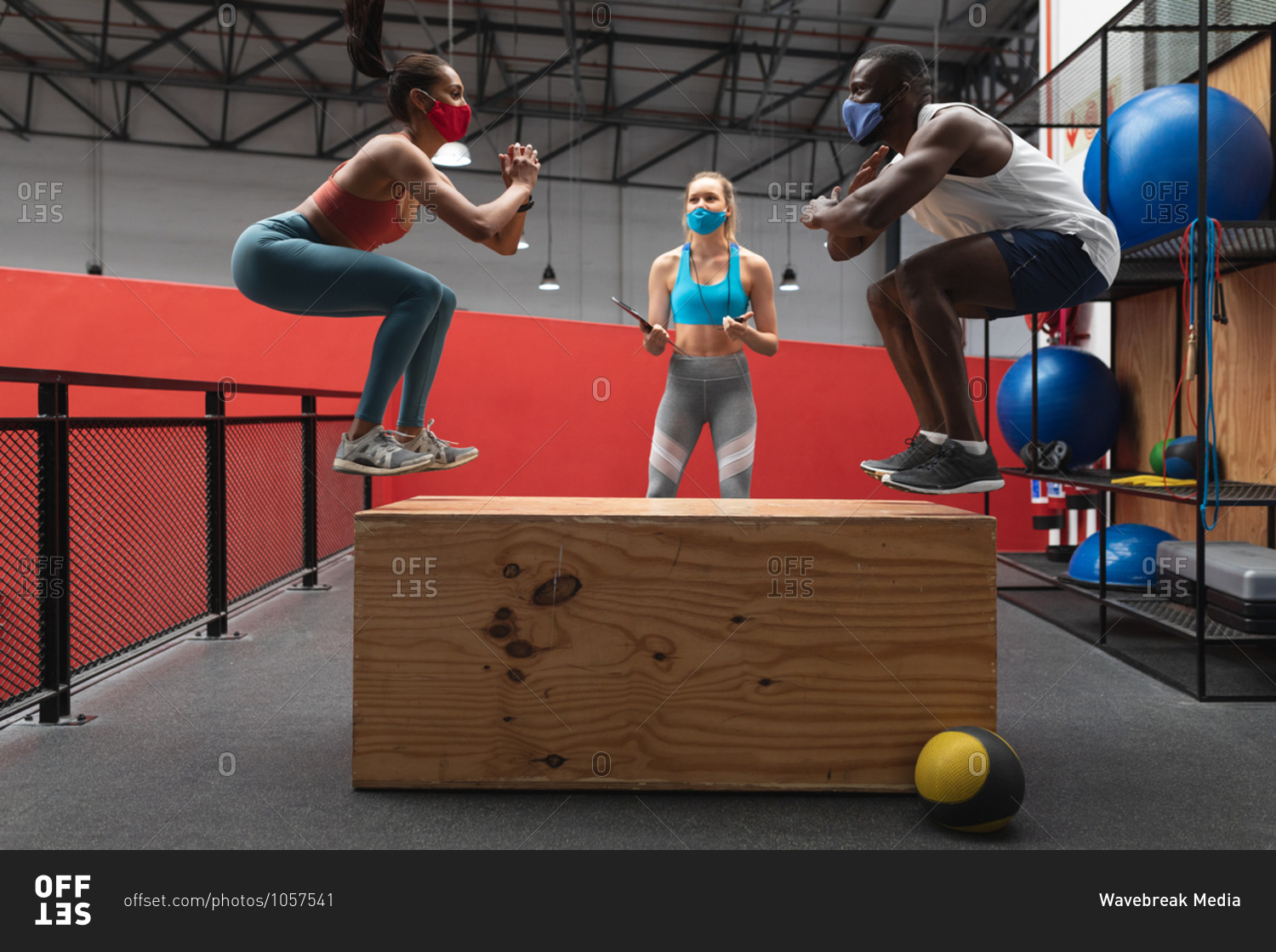 Fit African American man and Fit caucasian woman wearing face masks jumping on wooden plyo box in the gym while caucasian female fitness trainer holding stopwatch and clipboard.