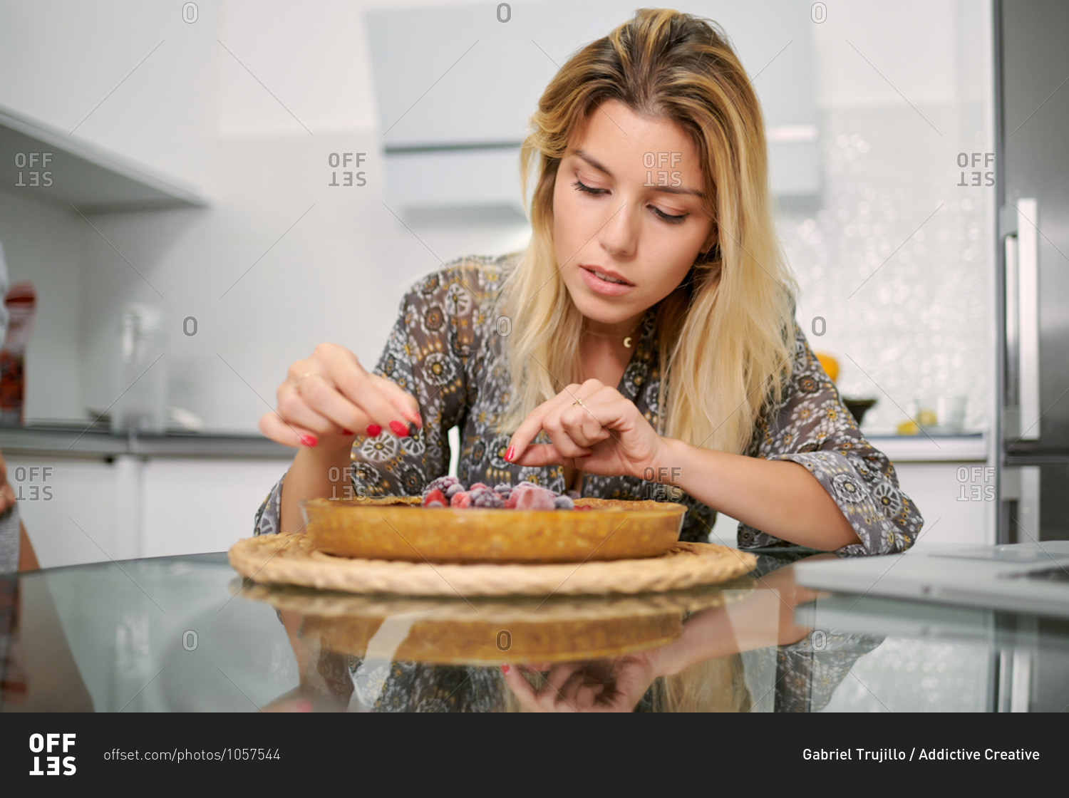 Focused young female garnishing homemade vegan tart with frozen berries while sitting at table with laptop in home kitchen