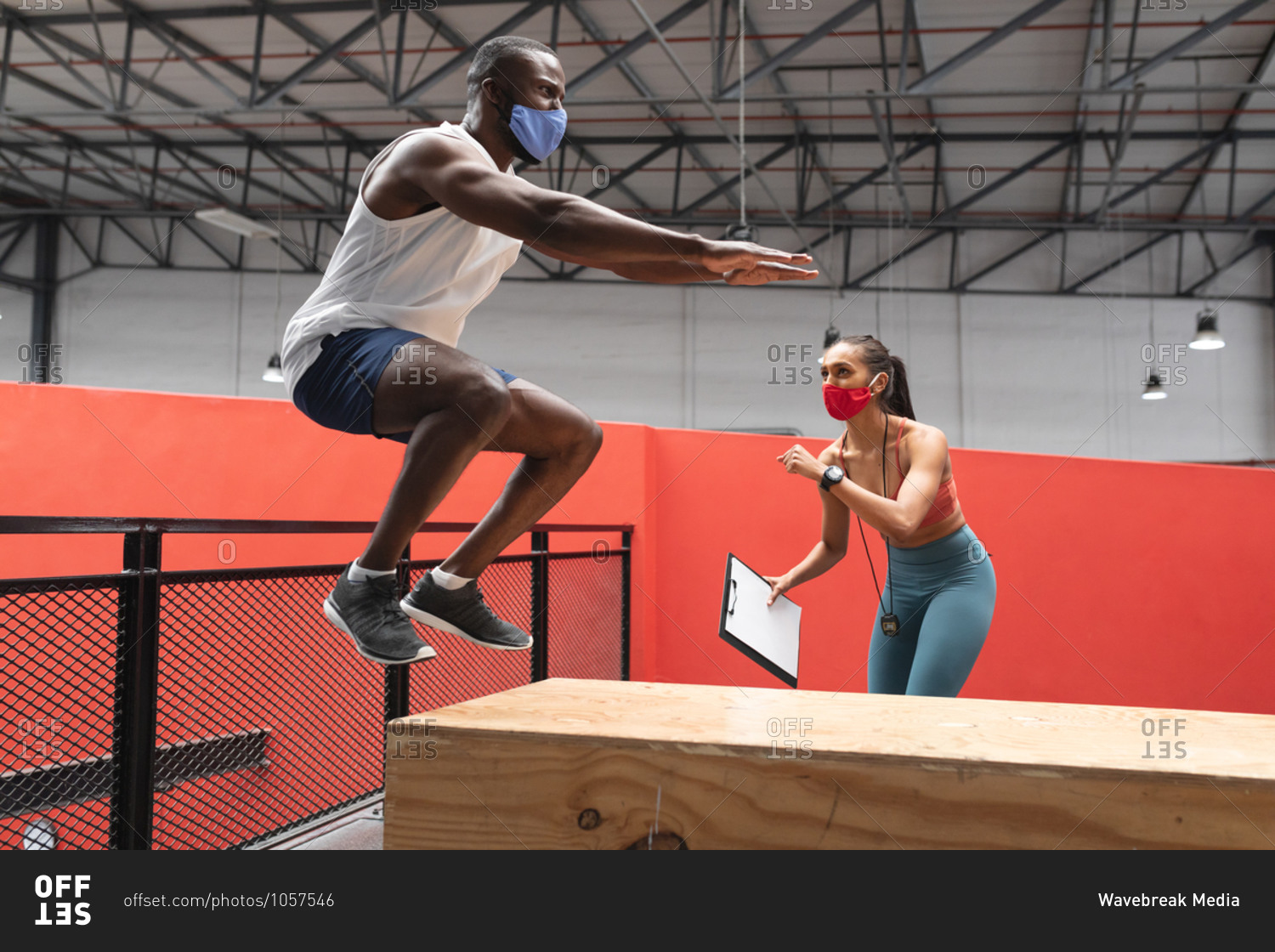 Fit African American man wearing face mask jumping on wooden plyo box in the gym while caucasian female fitness trainer holding stopwatch and clipboard.