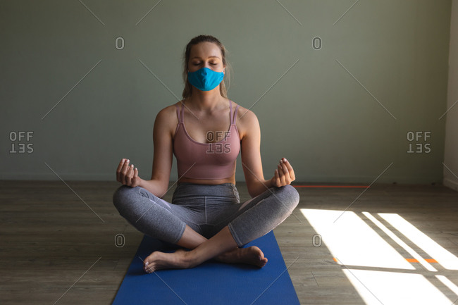 Fit caucasian woman wearing face mask practicing yoga while sitting on yoga mat in the gym.