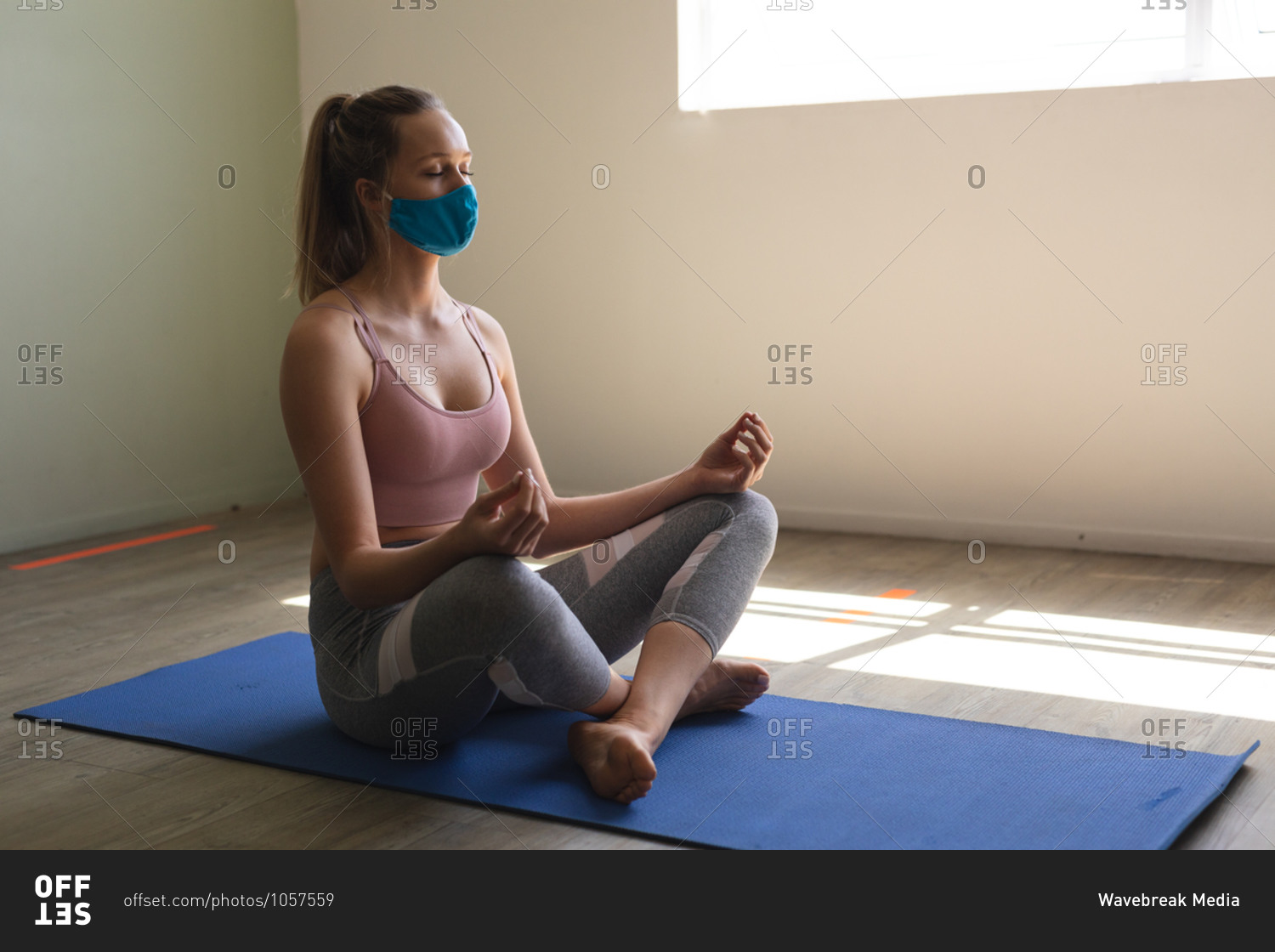 Fit woman caucasian wearing face mask practicing yoga while sitting on yoga mat in the gym.