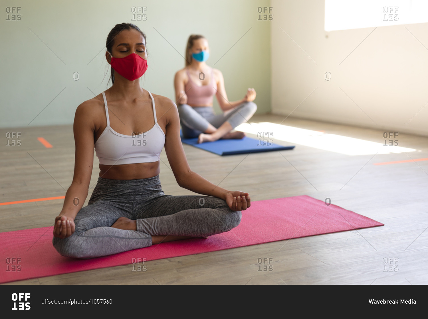 Two fit caucasian woman wearing face masks practicing yoga while sitting on yoga mats in the gym.