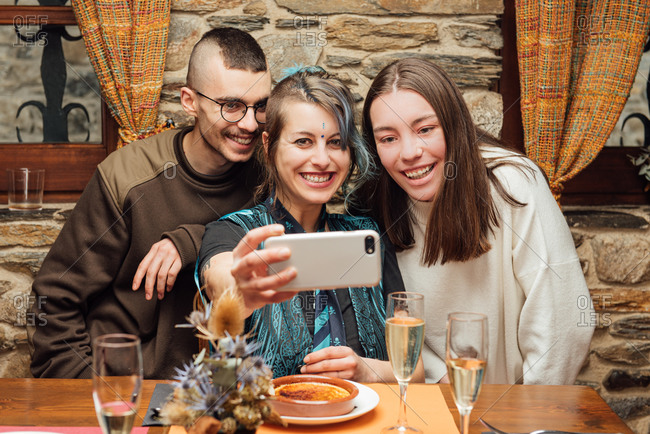 Delighted company of hipsters with fancy appearance sitting at table in cafe and taking selfie on smartphone while spending weekend together and having fun