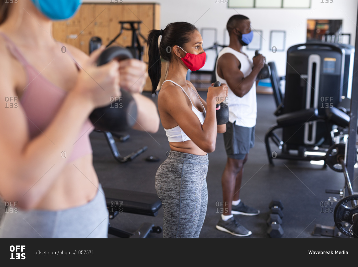 Fit African American man and two Fit caucasian woman wearing face masks performing exercise with kettlebells in the gym.
