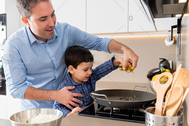 Glad young father teaching adorable little son to cook while pouring oil in pan together in modern light kitchen